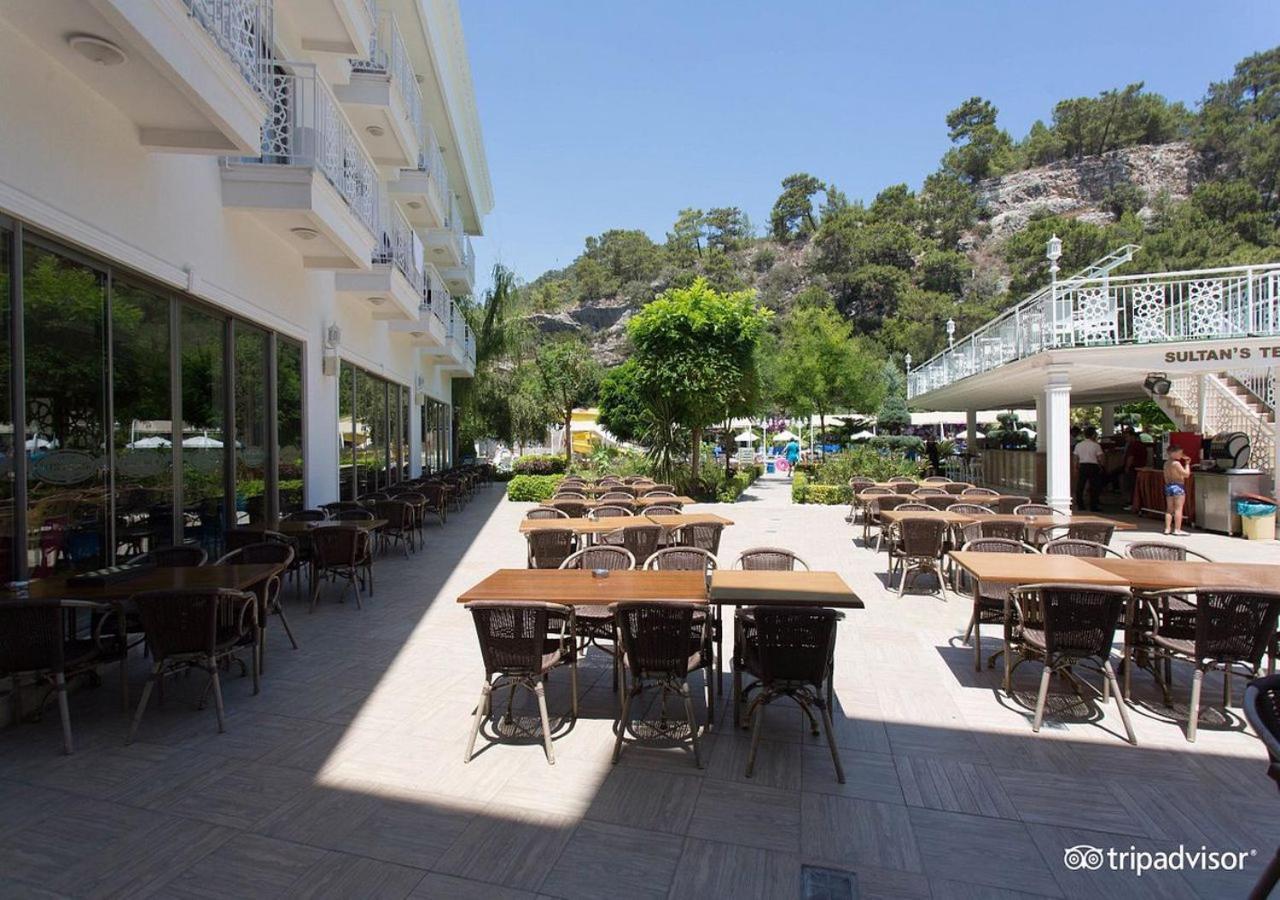 Miramor Hotel & Spa Antalya Extérieur photo The photo shows an outdoor dining area, likely at a restaurant or cafe. There are several tables with chairs arranged neatly on the paved ground, and some greenery, such as trees and plants, is visible in the background. The setting appears bright an